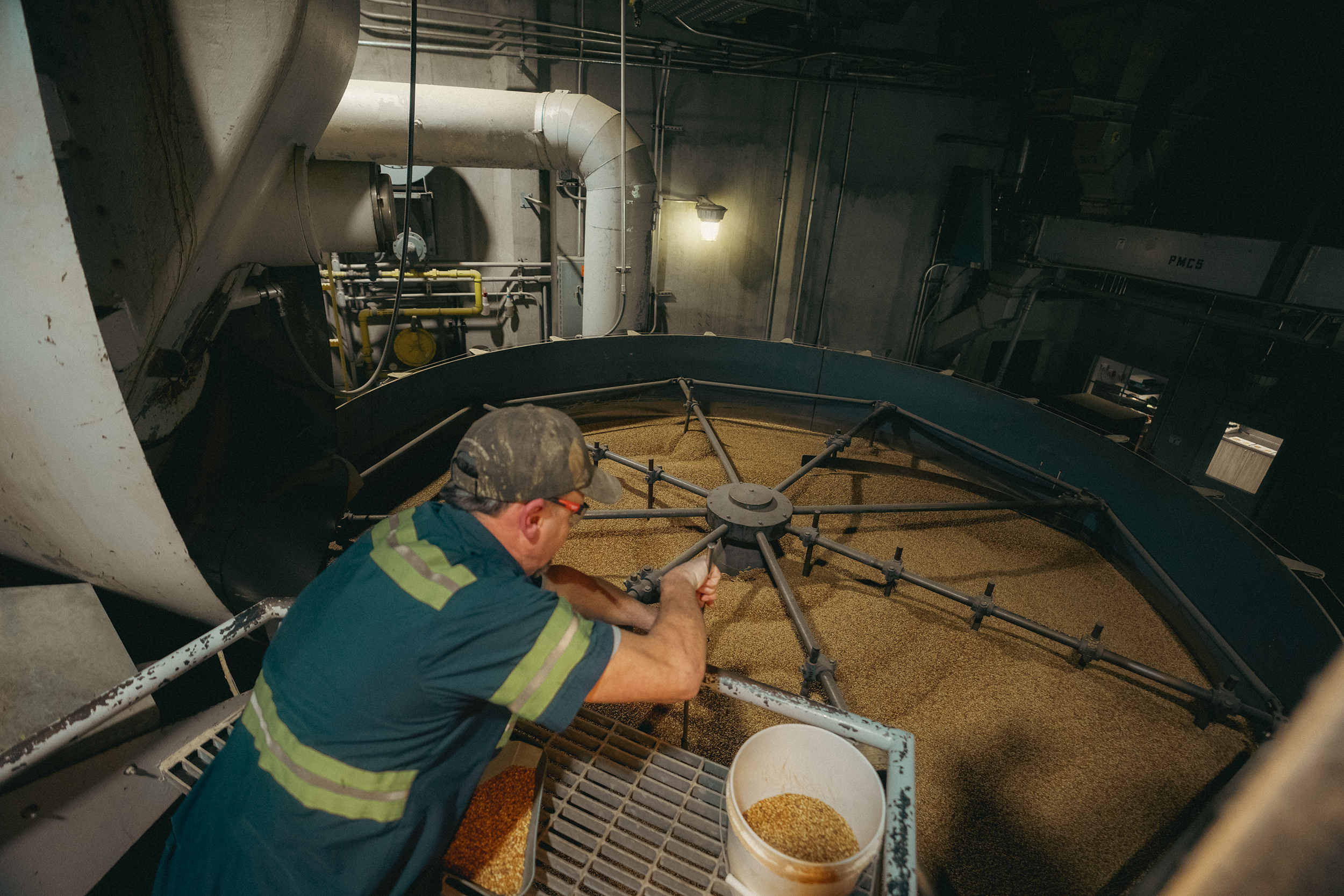 The top down view of a man in a safety reflective shirt using a rod to stir a large vat of grains.